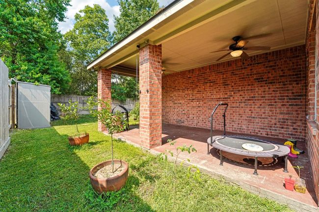 This photo showcases a cozy backyard with a covered patio featuring brick pillars, a ceiling fan, and a small trampoline. The yard is fenced, with grassy areas and potted plants, offering a pleasant outdoor space for relaxation or play. | Image 27