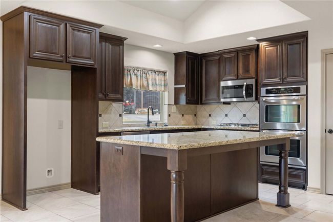 Kitchen featuring dark brown cabinetry, decorative backsplash, light stone counters, a center island, and stainless steel appliances | Image 8