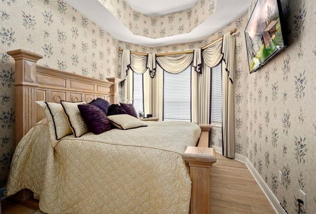 This is a traditional-style bedroom featuring floral wallpaper, a wooden headboard, elegant window draperies, and a matching bedspread with decorative pillows. The room has a classic charm and includes a flat screen T.V. on the wall. | Image 15