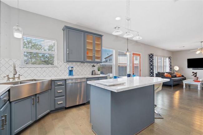 This photo showcases a bright, modern kitchen and living area. The kitchen features sleek blue cabinets, a stylish backsplash, stainless steel appliances, and a large island with pendant lighting. The open layout flows into a cozy living space with ample natural light and contemporary decor. | Image 18
