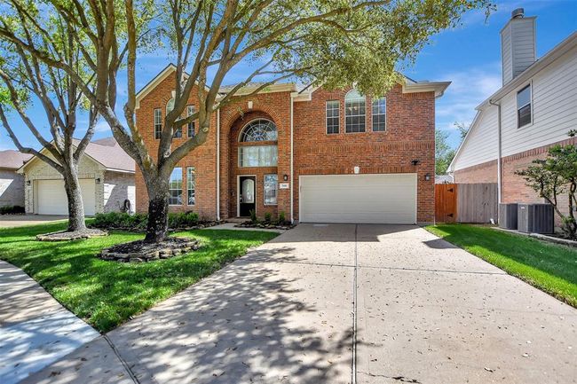 This photo shows a two-story brick home with a well-maintained front yard, featuring mature trees and a large driveway leading to a two-car garage. The house has a prominent arched window above the entrance, adding character to its facade. | Image 47
