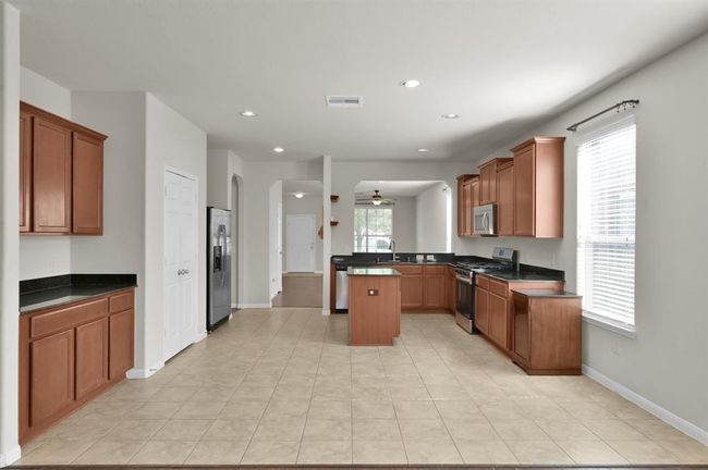 Kitchen with modern stainless steel appliances, granite countertops, and wooden cabinetry. It features tile flooring, an island, and an open layout that connects to a bright living area with hardwood floors. | Image 12