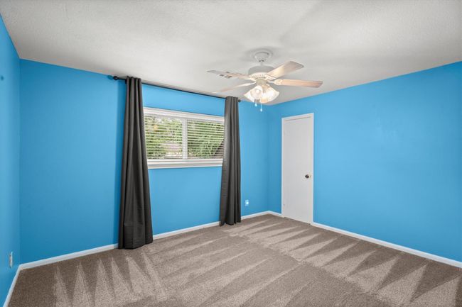 This secondary room features vibrant blue walls, a ceiling fan, and a large window, offering ample natural light. The carpet is clean and in good condition, creating a cozy atmosphere! | Image 31