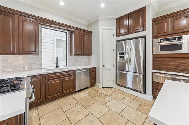 This kitchen features rich wooden cabinetry, stainless steel appliances, and a bright, neutral tile floor. It includes a built-in oven, a large refrigerator, and plenty of counter space for meal preparation. The design is both functional and stylish. | Image 11