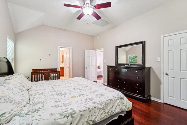 This spacious bedroom features warm wood flooring, a ceiling fan, and ample natural light. It includes a large bed, a dresser with a mirror, and a crib. The room connects to a bathroom and has neutral walls, offering a cozy and inviting atmosphere. | Image 17