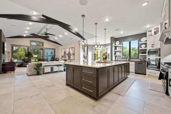 This photo showcases a modern, open-concept living space with high ceilings and exposed beams. The large kitchen features an expansive island with pendant lighting, sleek cabinetry, and high-end appliances. Adjacent is a bright living area with large windows offering plenty of natural light and views of greenery. The space blends contemporary design with warmth and functionality. | Image 18
