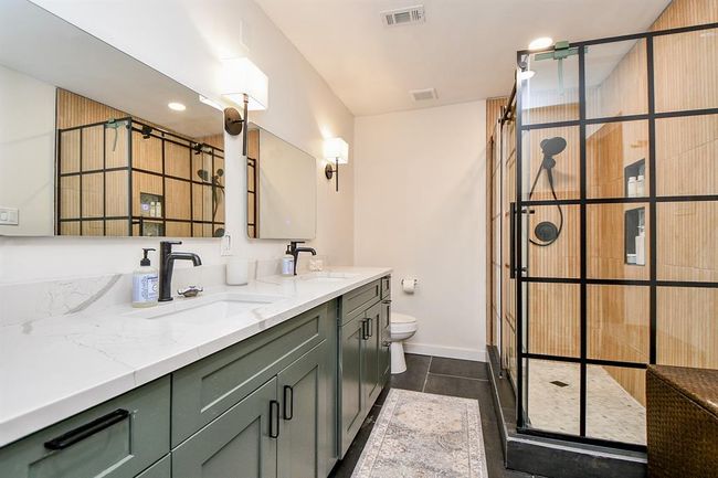 Modern bathroom featuring a double vanity with sleek black fixtures, a large mirror, and stylish lighting. The spacious shower has a glass enclosure with black grid accents and warm wood-like tiles. | Image 28