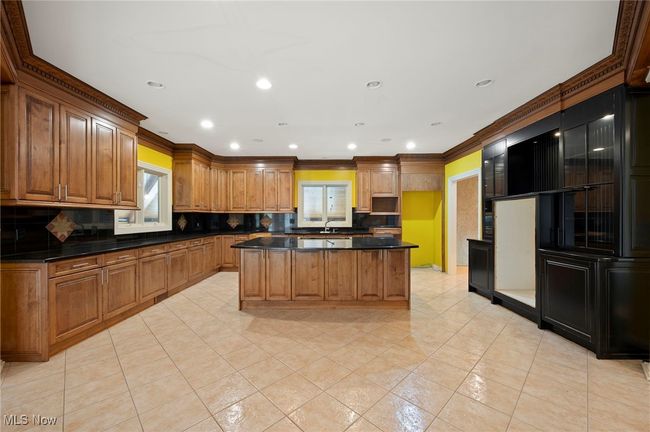 Kitchen featuring backsplash, dark countertops, light tile patterned flooring, crown molding, and brown cabinets | Image 15