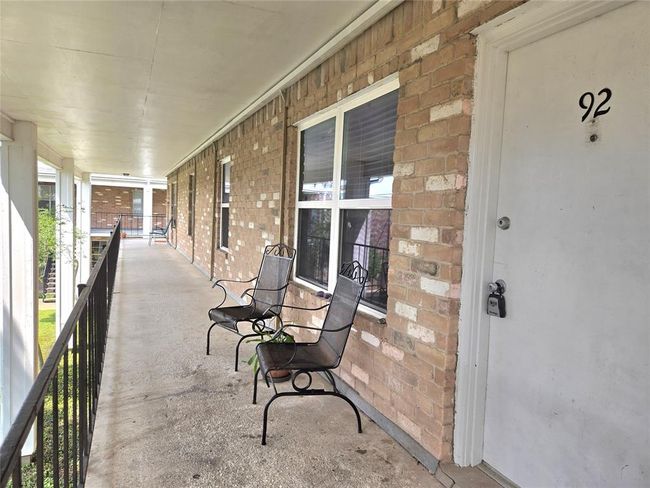 This photo shows a covered walkway leading to a unit entrance with a brick exterior. It features two metal chairs and has railing for safety. The area is well-lit and offers a view of the surrounding greenery. | Image 21