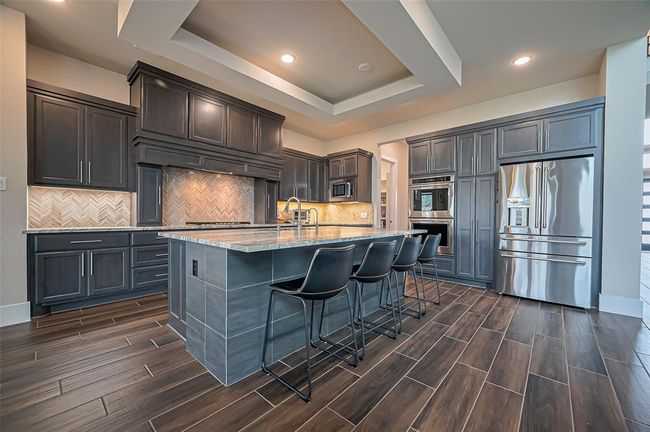 Modern kitchen with dark cabinetry and stainless steel appliances. Features a large island with seating, granite countertops, and a stylish herringbone backsplash. Bright, open space with elegant wood-look tile flooring. | Image 11