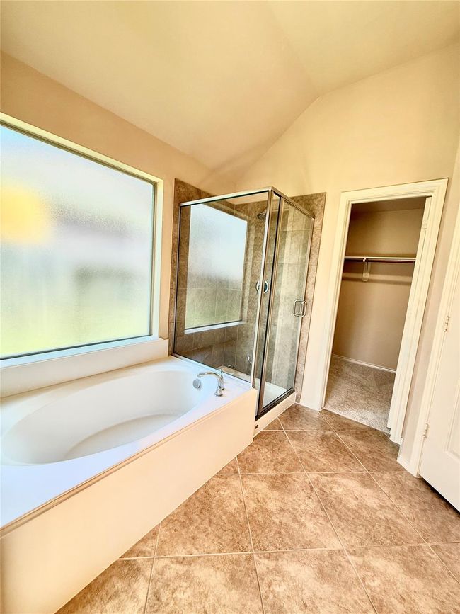 This bathroom features a spacious bathtub with a large frosted window for natural light, a glass-enclosed shower, and an adjoining walk-in closet. The floor is tiled, and the space has a high ceiling for an airy feel. | Image 16