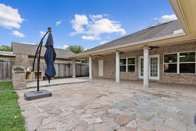 The photo shows a spacious, covered patio area with stone flooring, perfect for outdoor relaxation and entertainment. The patio includes a built-in brick fireplace and overlooks a fenced backyard. | Image 39