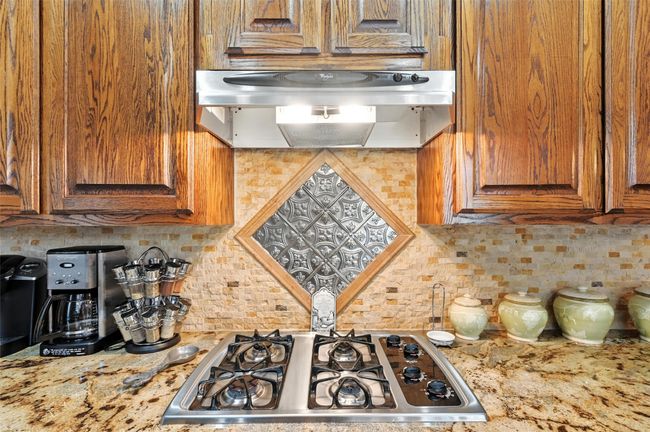 Kitchen featuring stainless steel gas stovetop, exhaust hood, backsplash, brown cabinetry, and light stone countertops | Image 7