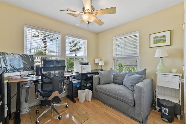 This cozy home office features large windows with plenty of natural light, a comfortable chair, and a spacious desk. The room includes a small couch, a ceiling fan, and light-colored walls, creating a bright and inviting workspace. | Image 23