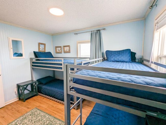 This bedroom features twin bunk beds with blue bedding, and natural light comes through two windows, complemented by a side table and decorative wall art. | Image 17