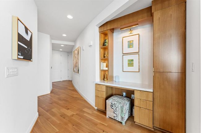 This photo showcases a modern hallway with built-in cabinetry featuring a small desk area. The space is well-lit with recessed lighting and accented with contemporary artwork, creating an inviting and functional entryway. | Image 6