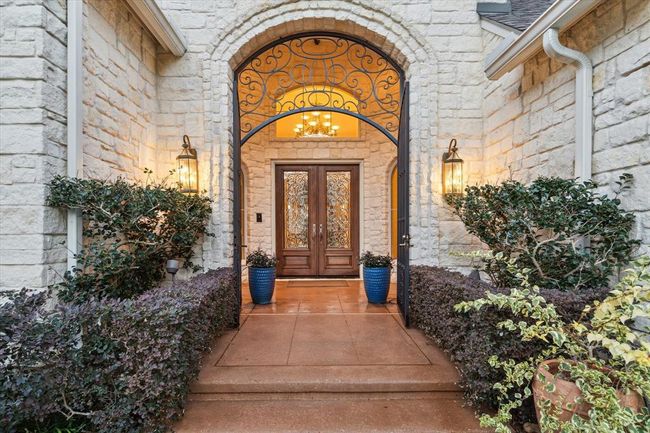 This inviting home entrance features a grand arched doorway with decorative wrought iron details, elegant double doors, and warm exterior lighting. Flanked by lush greenery and vibrant planters, it offers a welcoming and picturesque approach. | Image 6
