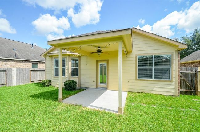 This photo shows a backyard with a small covered patio attached to the house. The patio has a ceiling fan and string lights, overlooking a grassy lawn enclosed by a wooden fence. The house features light-color siding and multiple windows, offering a cozy outdoor space. | Image 29