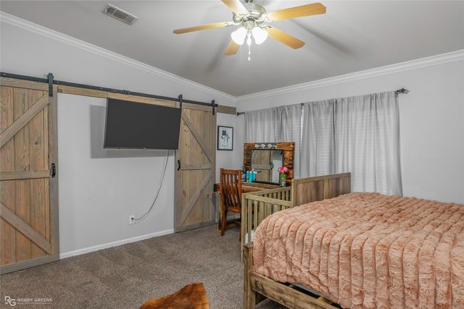 Carpeted bedroom with a barn door, ceiling fan, visible vents, baseboards, and ornamental molding | Image 24