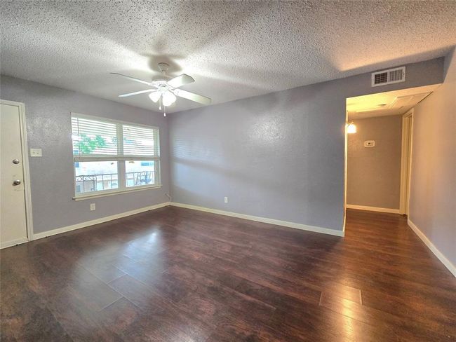 Bright living room with light purple walls, dark wood flooring, and a ceiling fan. Large window lets in natural light. Adjacent hallway leads to other parts of the home. | Image 6