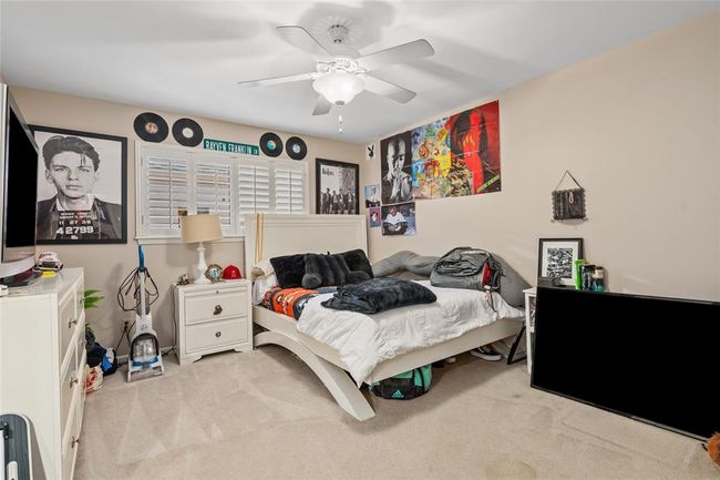 This bedroom features a neutral color scheme with soft carpeting and light-colored walls. A large window with plantation shutters allows natural light to brighten the space. The ceiling is equipped with a fan and light fixture for added comfort. The room offers ample wall space for personalization and decoration. | Image 29