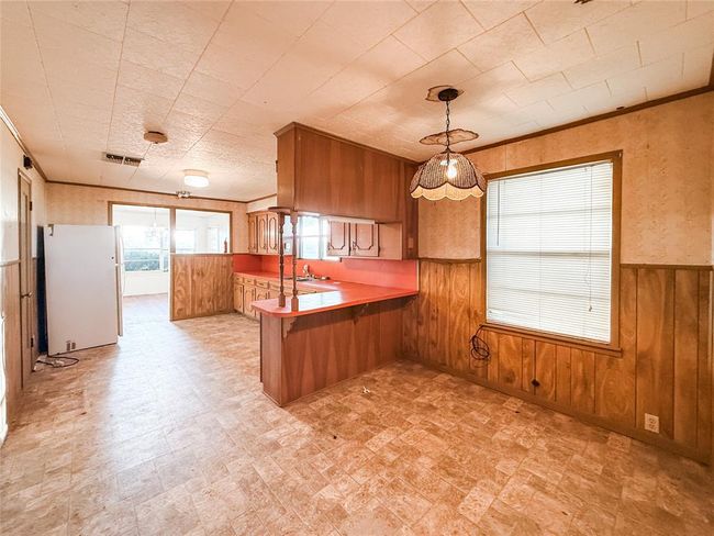 This photo shows a dated kitchen and dining area with wood paneling and a distinctive red countertop. There's a pendant light above the dining space and a visible refrigerator. The room has a spacious layout with linoleum flooring, offering potential for renovation. | Image 6