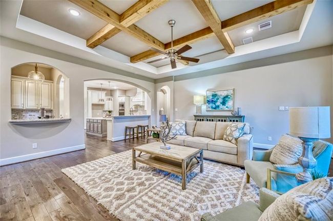 This spacious living area features a cozy, elegant design with exposed wooden beams and a tray ceiling. It flows seamlessly into the kitchen through wide archways, offering an open-concept feel. The room is well-lit, with modern furnishings and a plush area rug. Perfect for entertaining or relaxing. | Image 21