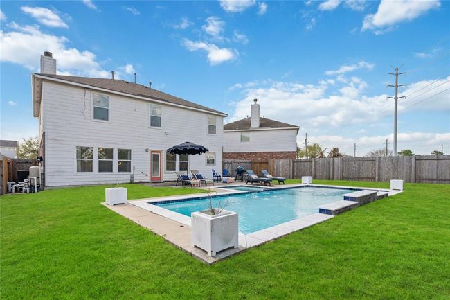 This photo showcases a spacious backyard featuring a large, inviting swimming pool surrounded by well-maintained green grass. The area includes lounge chairs for relaxation and is bordered by a wooden fence, offering privacy. The house exterior is visible, with multiple windows and a clean, white finish. | Image 8