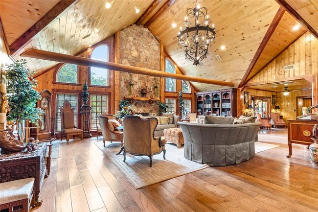 This spacious living room features a vaulted wood ceiling with exposed beams, a large stone fireplace, and abundant natural light from tall windows. It has elegant furnishings, a chandelier, and warm wood accents for a cozy, inviting atmosphere. | Image 25