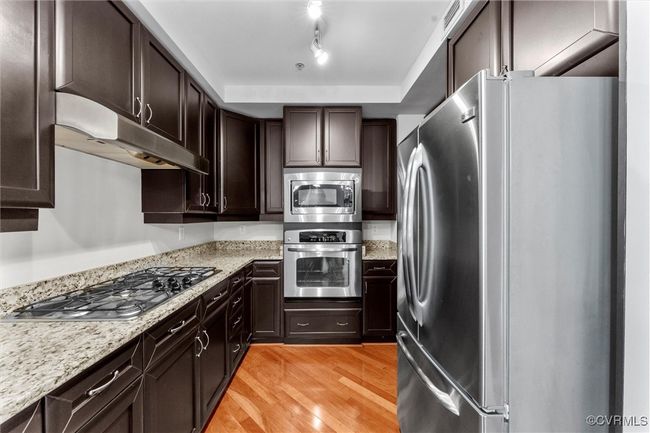 Kitchen featuring stainless steel appliances, light wood-style flooring, dark brown cabinets, light stone countertops, and under cabinet range hood | Image 10