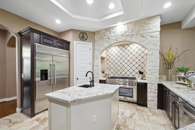This kitchen features a modern design with stainless steel appliances, a large double-door refrigerator, and a central island with a sink. It has stone accents around the stove, ample counter space, and a warm, inviting color palette. | Image 7