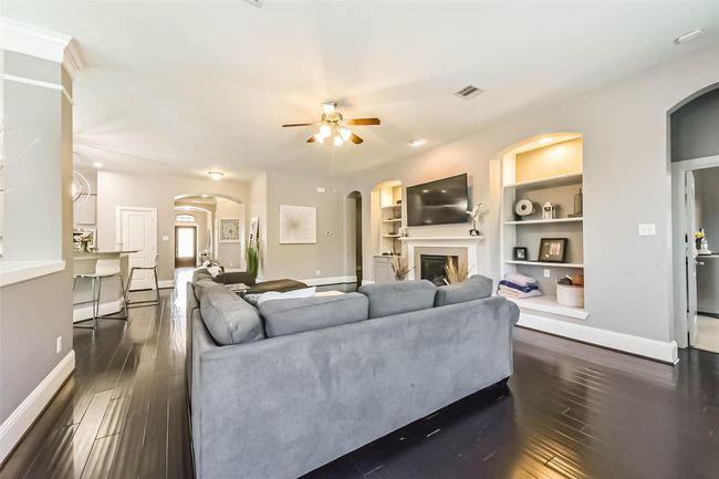 The inviting living room features dark hardwood floors, a cozy fireplace, and built-in shelves. A ceiling fan and recessed lighting add comfort and style. The open layout flows into the kitchen area, enhancing the spacious feel. | Image 10
