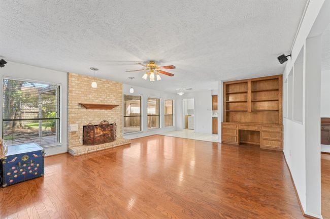 Spacious living room features a warm wooden floor, a cozy brick fireplace, built-in shelving, and large windows offering plenty of natural light. A ceiling fan and open layout create an inviting atmosphere, with easy access to the adjacent kitchen. | Image 5
