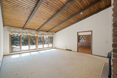 The large living room features a neutral palette, wood beamed vaulted ceilings, brick wall with fireplace and a wall of windows overlooking the spacious deck and backyard. | Image 2