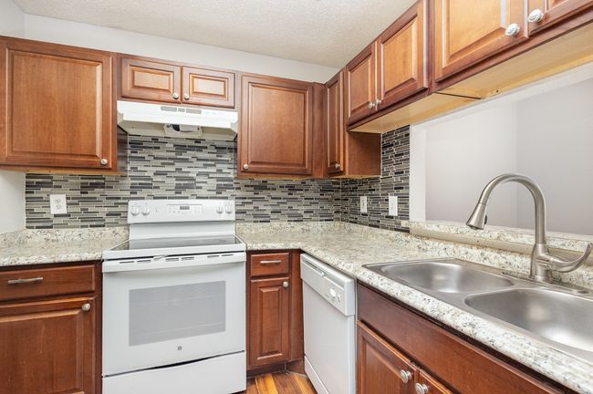 Kitchen featuring white appliances, backsplash, under cabinet range hood, a textured ceiling, and brown cabinetry | Image 7