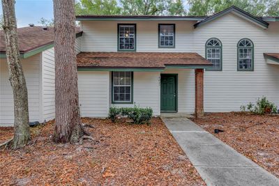 The image shows the exterior of a two-story townhouse with white siding and a brown shingled roof. The entrance is centered, featuring a green door with a small concrete pathway leading up to it. The windows on the second floor have a distinct design, with two tall, arched windows on the right and rectangular windows on either side of the front door. A large tree with a thick trunk is on the left side of the yard, which is covered in dry leaves and mulch with some low green shrubs near the house. The adjacent unit, partially visible on the right, has a similar design, and an ADT security sign is placed near its entrance. The sky is blue, and the area is surrounded by leafy trees. | Image 1