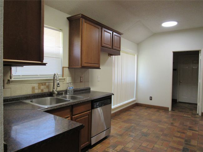 Kitchen featuring dark countertops, dishwasher, a sink, brick floor, and baseboards | Image 5