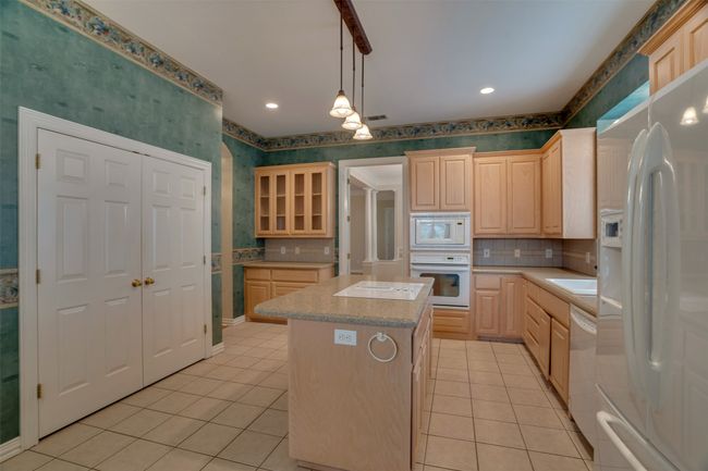 Kitchen with white appliances, wallpapered walls, light brown cabinetry, light tile patterned floors, and a kitchen island | Image 12