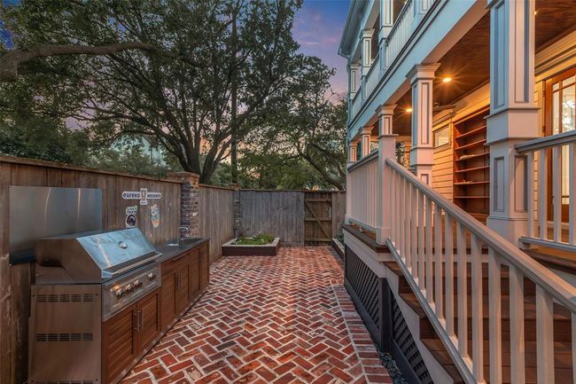 Charming outdoor kitchen with a built-in stainless steel grill, ideal for entertaining. The area is bordered by a wooden fence and lush trees, providing privacy and a serene atmosphere. A cozy porch with elegant railings adds to the inviting ambiance. | Image 38