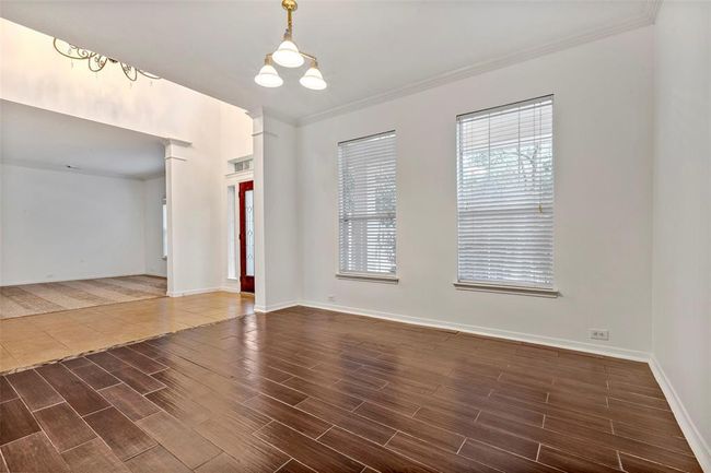 This photo showcases a bright, airy formal dining room with wood like tile flooring. It features large windows with blinds, a chandelier, and an open layout leading to another room. | Image 17