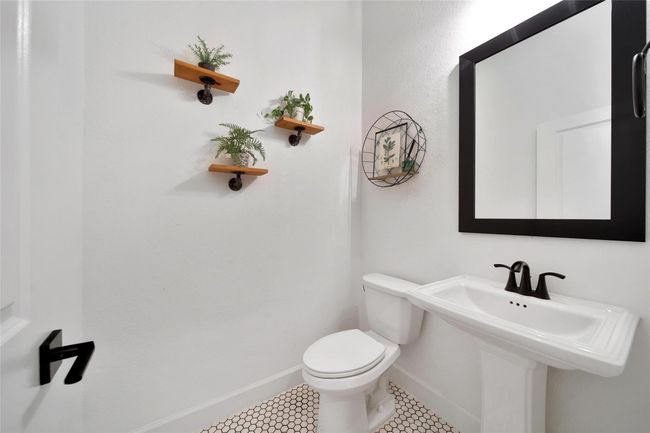 This is a modern, minimalist bathroom with a pedestal sink and toilet. It features a large black-framed mirror, white walls, and patterned floor tiles. Small decorative shelves with plants add a touch of greenery. | Image 40