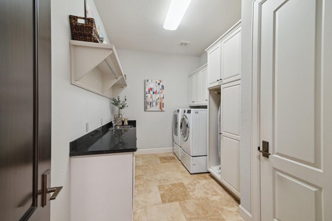 This light-filled laundry room, conveniently located just off the kitchen, is thoughtfully crafted for both efficiency and ease. Abundant cabinetry ensures plenty of storage, while polished granite countertops create a practical surface for folding and organizing. A deep utility sink, integrated hanging rods, and overhead shelving maximize utility, while the neutral tones and travertine flooring lend a crisp, refined finish to this everyday space. | Image 39