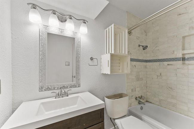 This bathroom features a sleek vanity with a decorative mirror and modern lighting. The shower-tub combo is adorned with elegant tile work, including a stylish accent strip. Ample storage is provided by wooden shelves, adding both functionality and charm. | Image 18