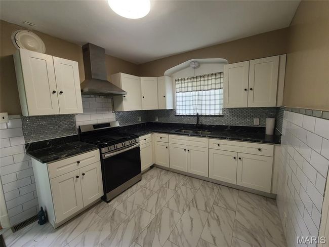 Kitchen with stainless steel range with gas cooktop, wall chimney exhaust hood, light marble finish floors, white cabinets, and dark stone countertops | Image 13