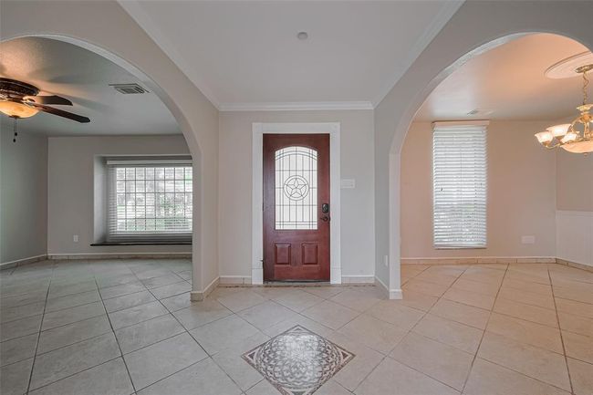 Step into a welcoming foyer that showcases elegant archways, neutral tile flooring, and a rich wood front door with decorative tile inlay. Crown molding and natural light set the tone for the home’s warm, inviting aesthetic. | Image 6