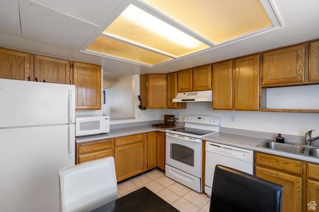 Kitchen with white appliances, light tile patterned floors, under cabinet range hood, brown cabinets, and a sink | Image 9