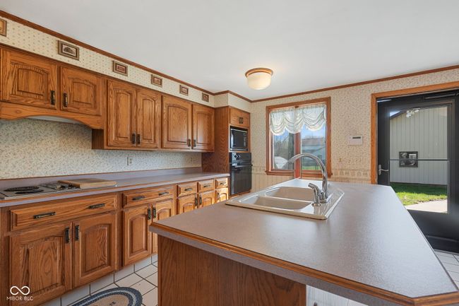 kitchen with a sink, black appliances, wallpapered walls, brown cabinets, and a kitchen island with sink | Image 14