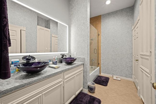 This is a modern bathroom featuring a double vanity with stylish vessel sinks, ample cabinet space, a large mirror with backlighting, and a walk-in shower with glass enclosure. The space is accented with coordinated dark towels and bath mats, giving it a cohesive and elegant look. | Image 28