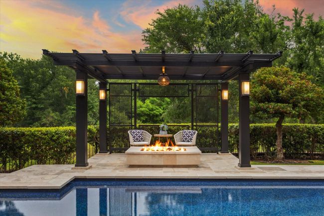 Situated amid vibrant landscaping and a canopy of trees along the creek, this modern pergola with tranquil slatted wood panels sets the stage for a cozy fireside lounge—ideal for relaxed evening chats or roasting marshmallows with loved ones. A refined outdoor chandelier and ambient sconce lighting bathe the space in a soft, welcoming glow, creating a peaceful, light-filled retreat by the pool. The possibilities are truly endless in this resort-styled backyard! | Image 44