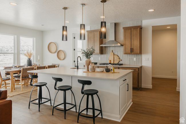 Kitchen with wall chimney exhaust hood, a breakfast bar area, tasteful backsplash, light countertops, and an island with sink | Image 4